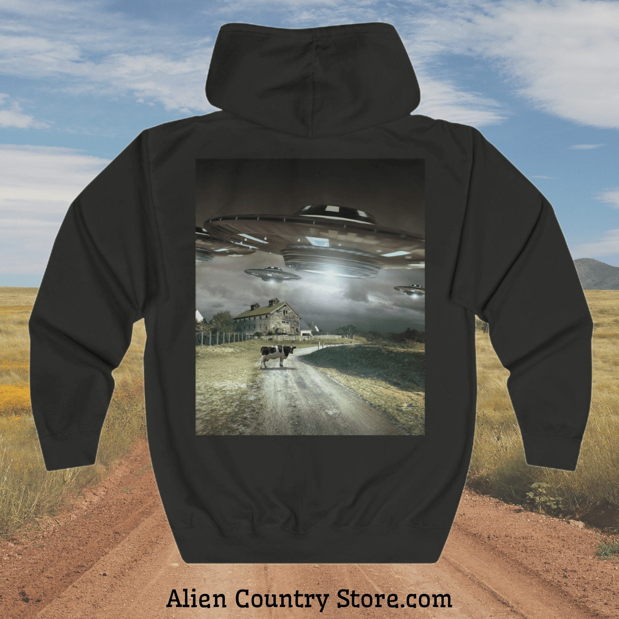 a photo of a hoodie with the image on the back. The image is a realistic pic of a cow standing in a country dirt road at sunset under a cloudy sky. Three UFO's hover nearby overhead.